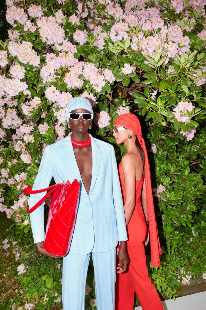 Two models in stylish blue and red outfits pose in front of a lush flower bush, showcasing a modern fashion aesthetic and color contrast.
