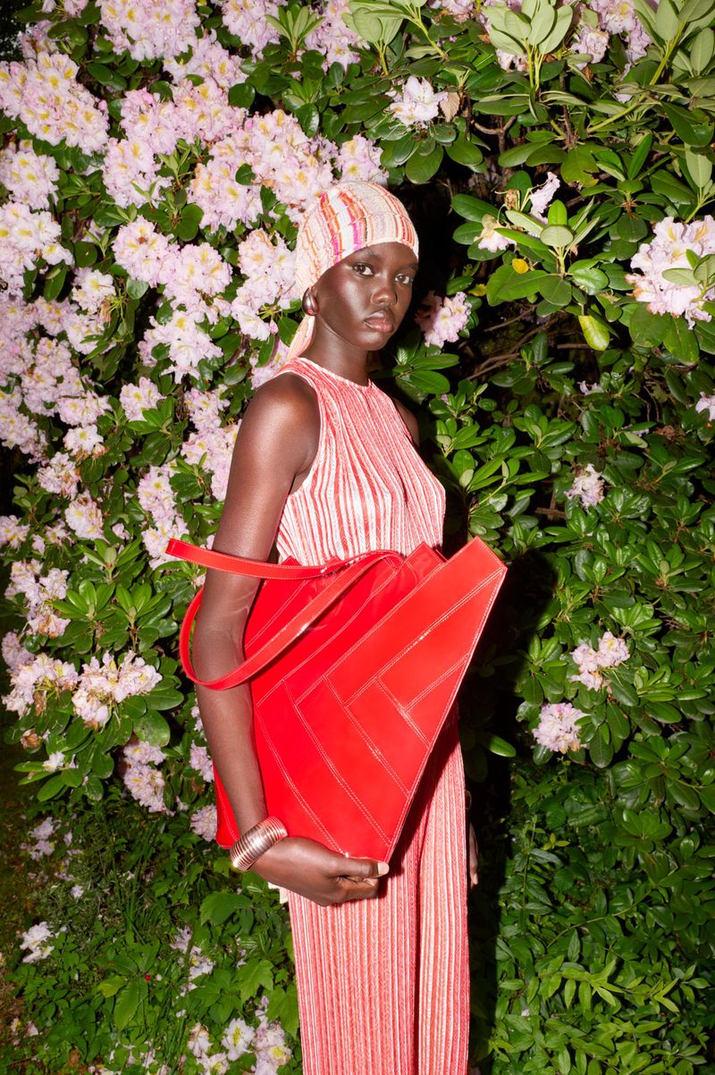 An adult woman stands outdoors wearing a red and white outfit, holding a large red bag, surrounded by green leaves and light pink flowers.