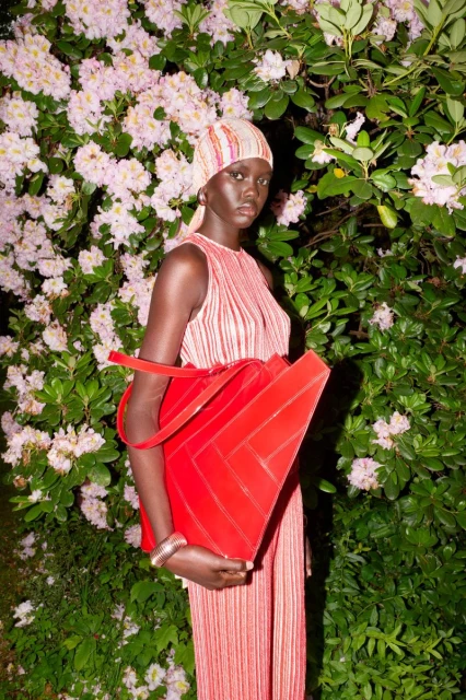 An adult woman stands outdoors wearing a red and white outfit, holding a large red bag, surrounded by green leaves and light pink flowers.