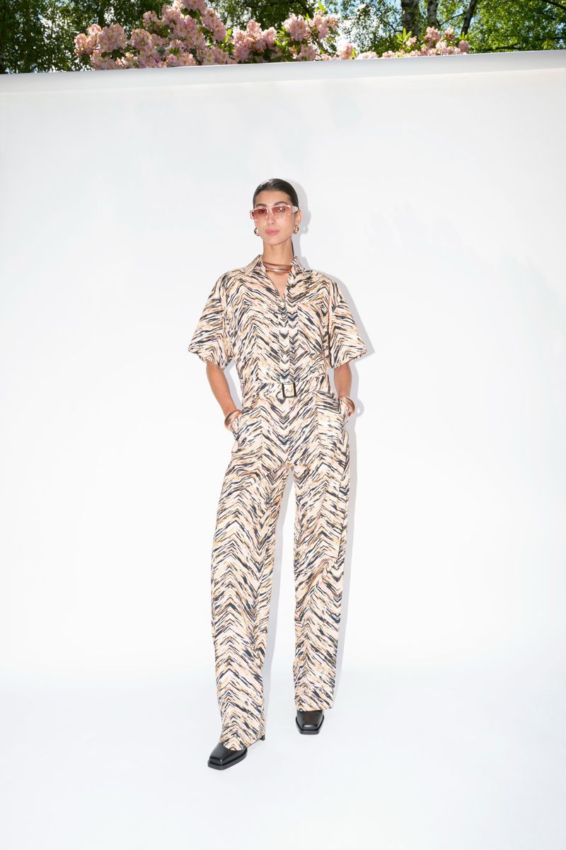 A woman in a patterned jumpsuit poses outdoors against a white backdrop with flowering trees visible above, creating a fashion-forward image.