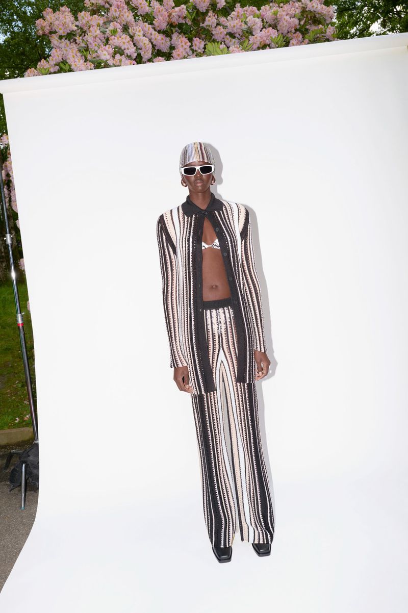Woman in a brown and white striped jacket and pants, accessorized with white sunglasses and a matching headscarf, poses in a studio.