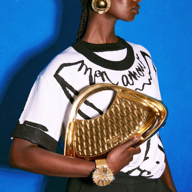 Dark-skinned woman with black-and-white sweater, gold handbag and jewelry poses against solid blue background in a fashion setting.