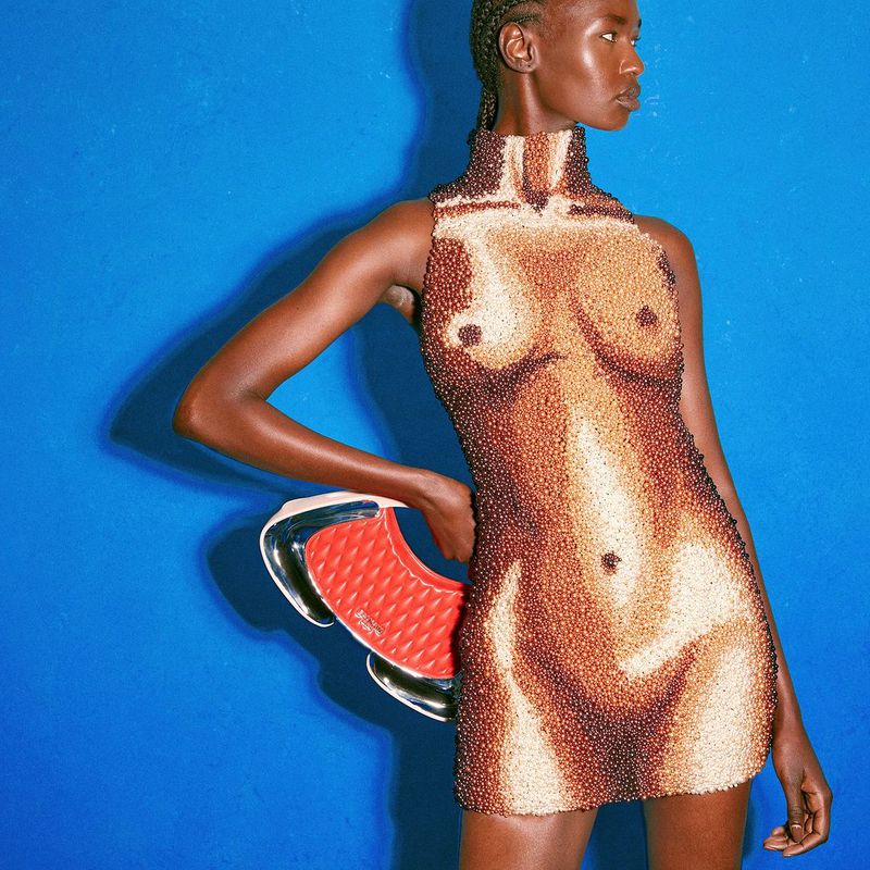 A fashion model in a beaded dress with a nude body print, holding a red handbag, poses against a vibrant blue backdrop in a studio setting.