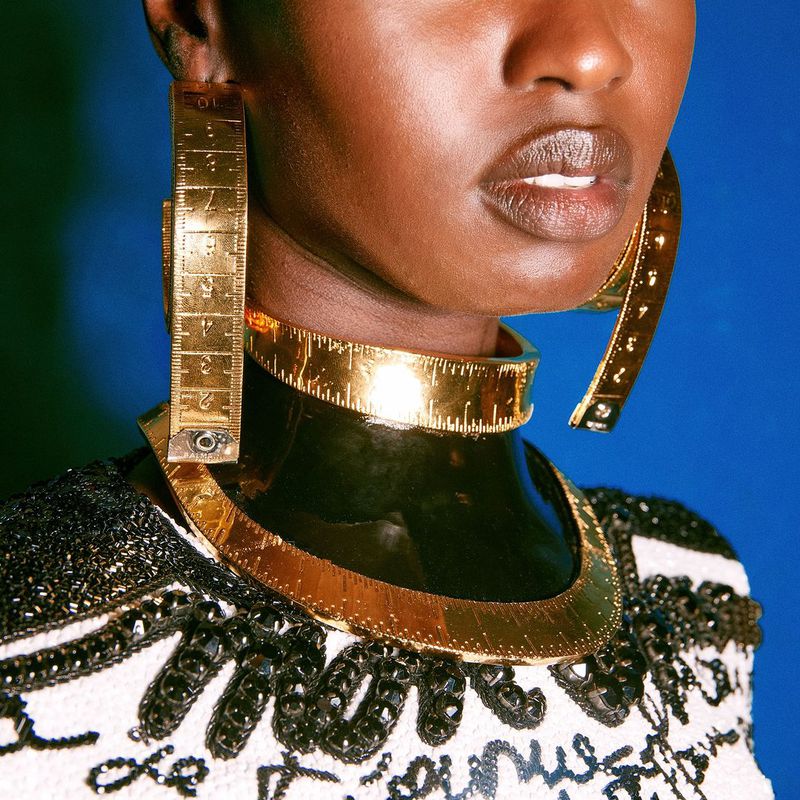 Close-up of a stylish Black woman wearing unique gold jewellery, including earrings and a choker that resemble measuring tapes against a blue background.