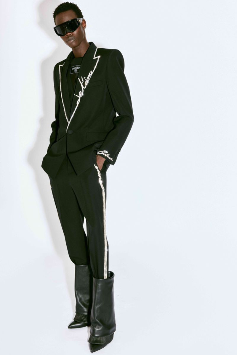 A Black fashion model wearing a black suit with white trim and black boots stands in front of a white background, studio lighting.