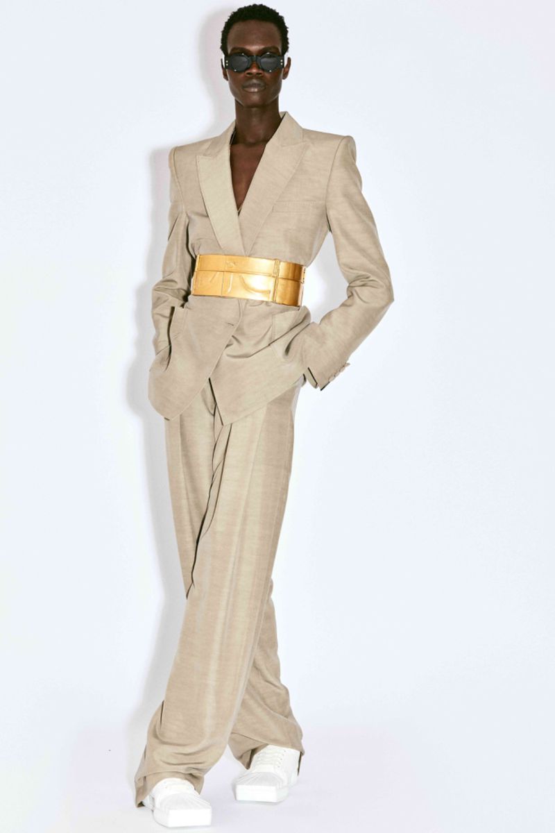 An androgynous fashion model poses in a tan blazer suit with a wide gold belt, sunglasses and white shoes against a white background.