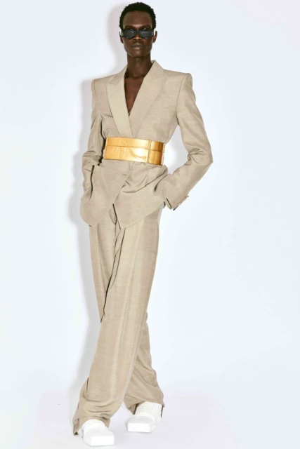 An androgynous fashion model poses in a tan blazer suit with a wide gold belt, sunglasses and white shoes against a white background.