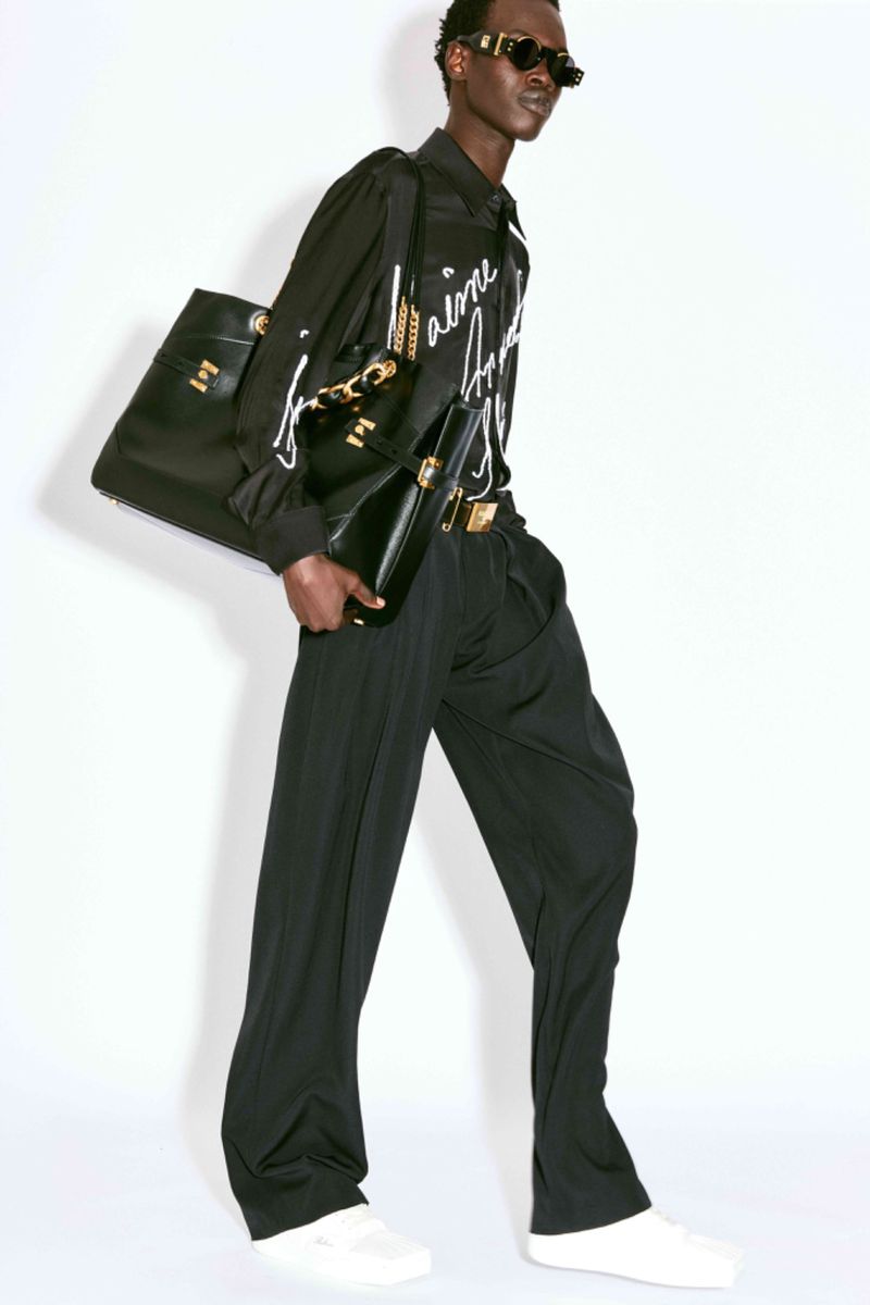 A slender Black male fashion model poses in a black printed shirt, black pants, white sneakers, and gold-framed sunglasses with purses.
