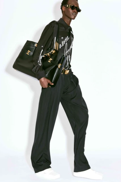 A slender Black male fashion model poses in a black printed shirt, black pants, white sneakers, and gold-framed sunglasses with purses.