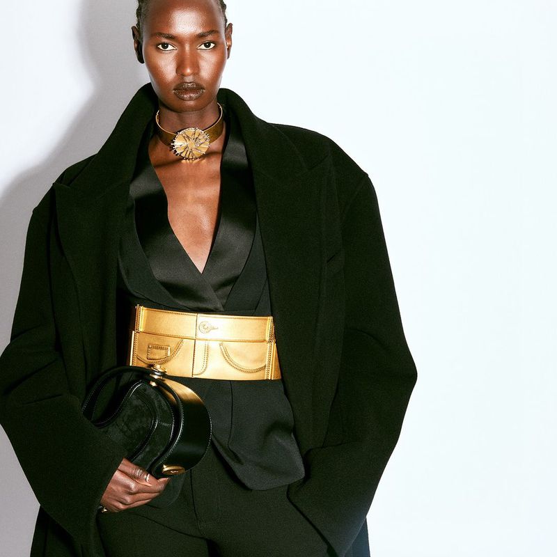 Elegant woman in black coat accessorized with gold collar, belt, and black purse poses against white backdrop in a fashion editorial.