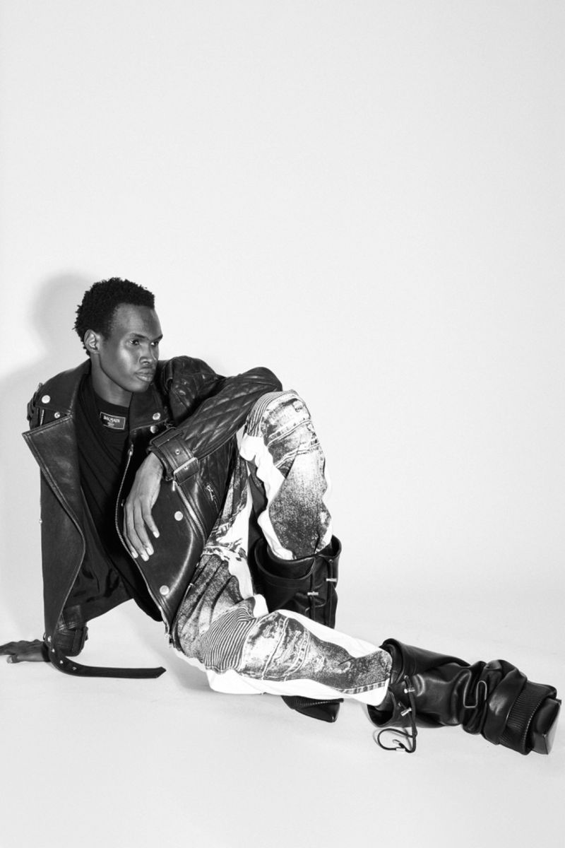 Monochrome fashion shot of a Black male model sitting on a white surface in a black leather jacket and printed pants in a photo studio.