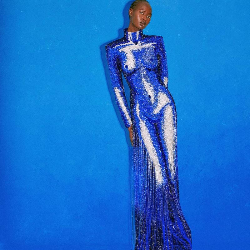 Elegant African woman poses in a sparkling blue bodycon gown with fringe against a solid blue backdrop for a fashion editorial shot.