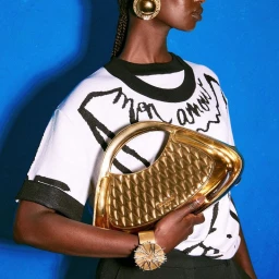 Model holding a metallic gold handbag, wearing a white graphic t-shirt and gold statement jewelry against a vibrant blue backdrop