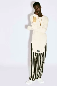 Black man wearing a cream ribbed sweater and black-and-white striped trousers against a white seamless background