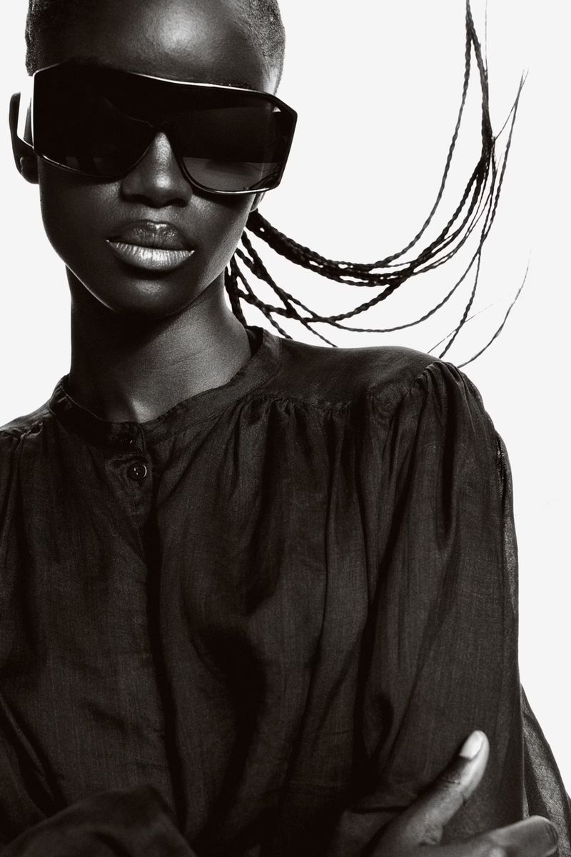 Portrait of a Black woman with sunglasses, wearing a dark blouse, against a white background, showcasing fashion and beauty elements.