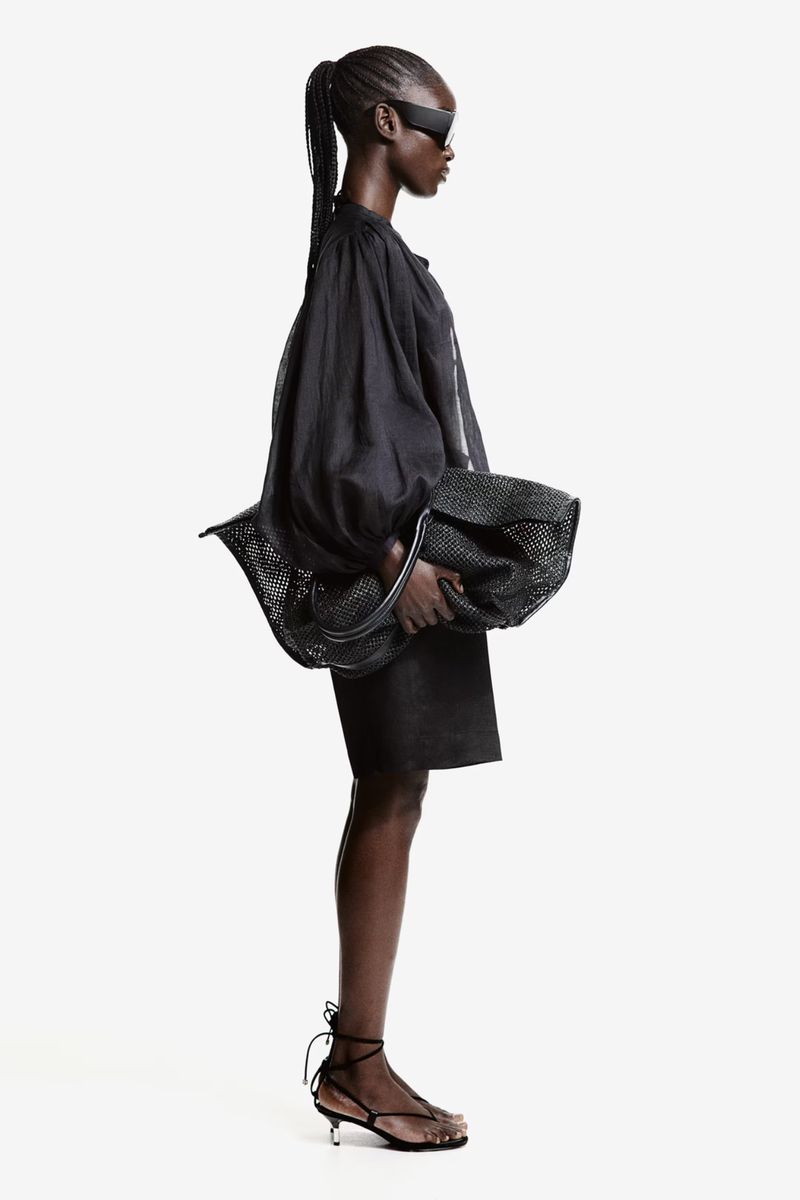 Profile of a black female fashion model with sunglasses wearing a black oversized top and skirt, holding a black mesh bag in a studio.