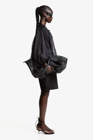 Profile of a black female fashion model with sunglasses wearing a black oversized top and skirt, holding a black mesh bag in a studio.