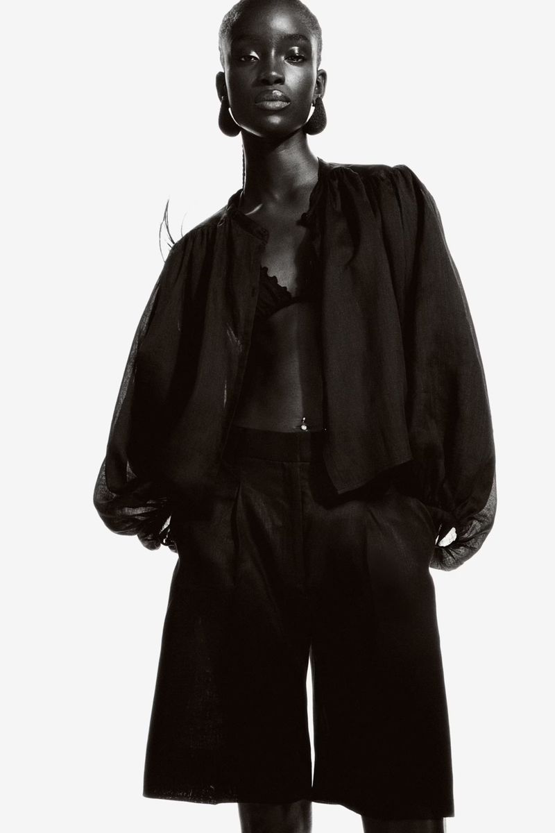 Elegant black and white fashion shot of a Black woman wearing a blouse, bralette, and high-waisted shorts against a clean white backdrop.