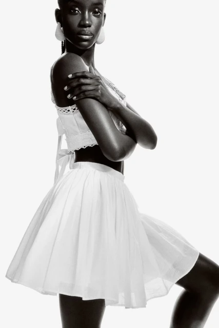 Monochrome fashion portrait of a black woman wearing a white cotton top and skirt with arms crossed in a photo studio setting.