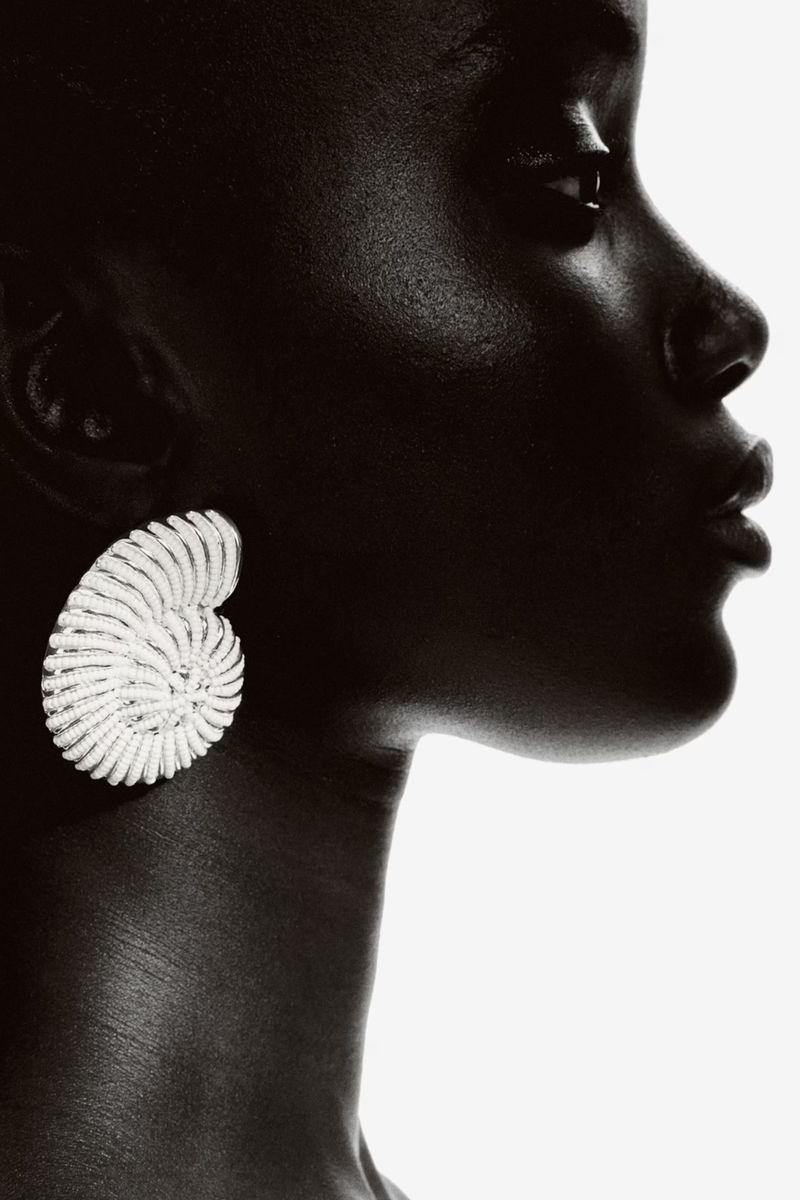 Close-up profile portrait of an adult woman with dark skin wearing a spiral-shaped earring against a white background.