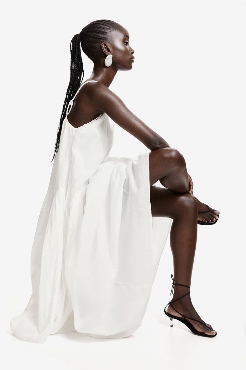 Model wearing a white dress and black strappy sandals seated against a white background for H&M