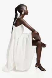 Model wearing a white dress and black strappy sandals seated against a white background for H&M