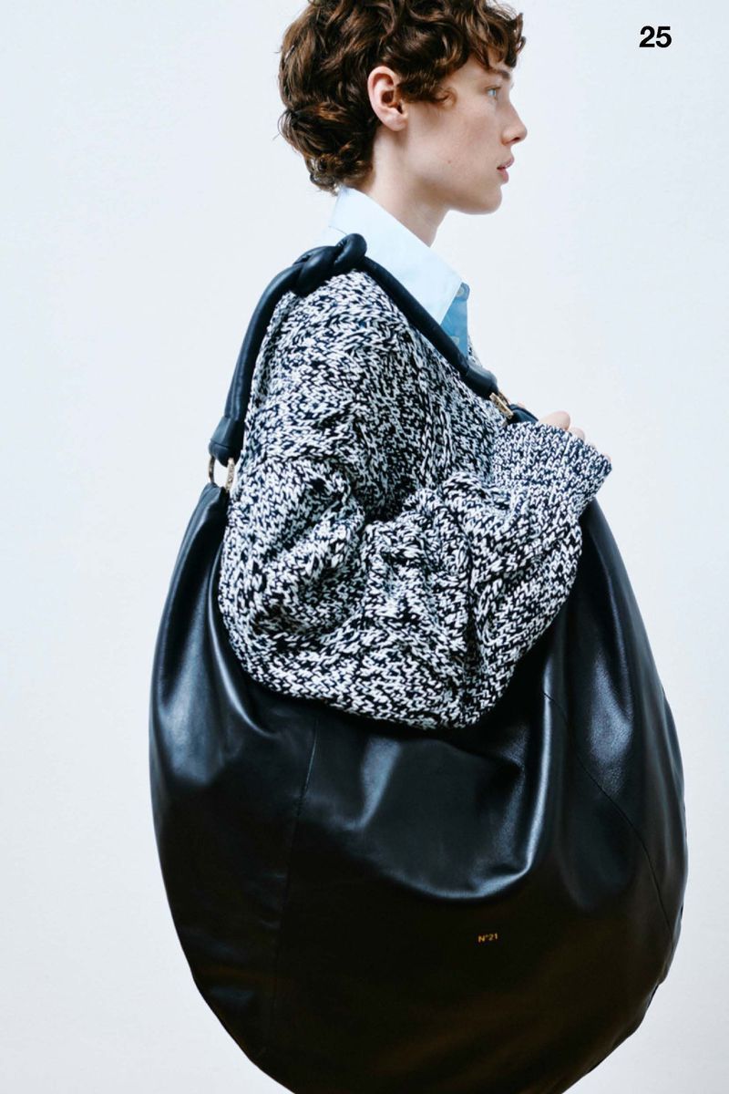 Fashion shot of a person with curly hair wearing a sweater and carrying a large black leather hobo bag over their shoulder in a photo studio.