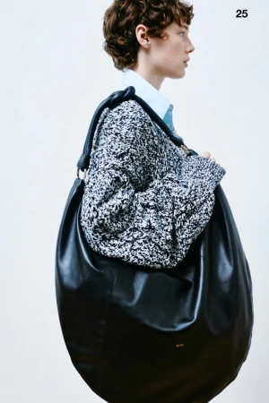 Fashion shot of a person with curly hair wearing a sweater and carrying a large black leather hobo bag over their shoulder in a photo studio.