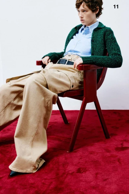 A young woman with short hair sits in a red chair wearing a green sweater and baggy beige pants on a red carpet against a white wall.