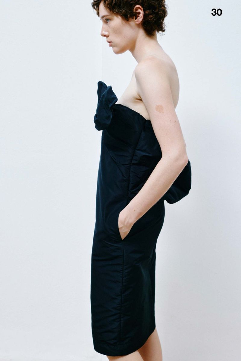 Profile view of a person in a dark blue, draped, strapless dress against a white background, featuring an upper-arm-length design.