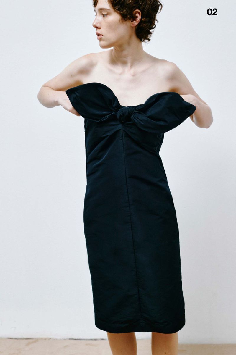 Adult female poses indoors wearing a black strapless dress with a large bow detail, looking off-camera in a fashion setting with studio lighting.