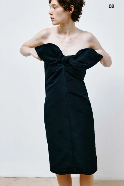 Adult female poses indoors wearing a black strapless dress with a large bow detail, looking off-camera in a fashion setting with studio lighting.