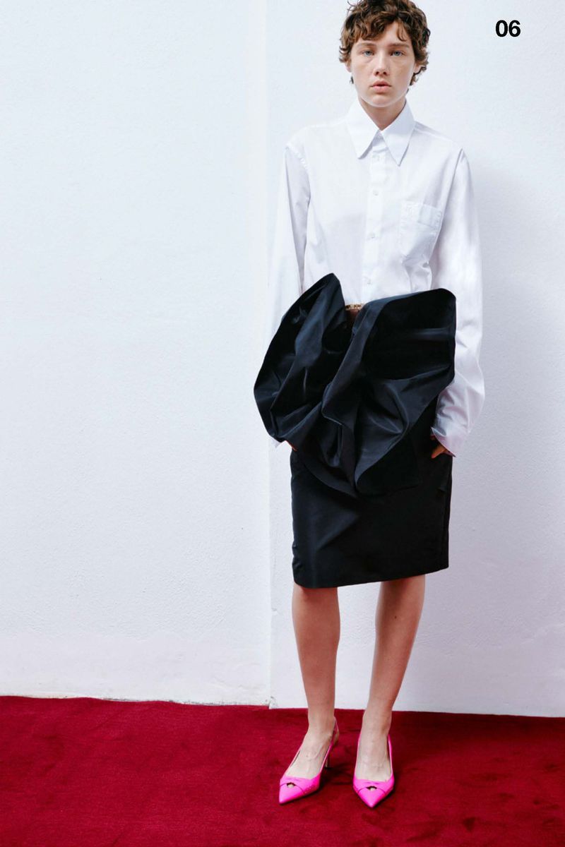 Fashion shot of a woman with short brown hair wearing a white button-down shirt, black skirt with bow detail, and pink pointed heels.
