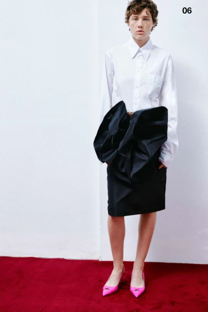 Fashion shot of a woman with short brown hair wearing a white button-down shirt, black skirt with bow detail, and pink pointed heels.