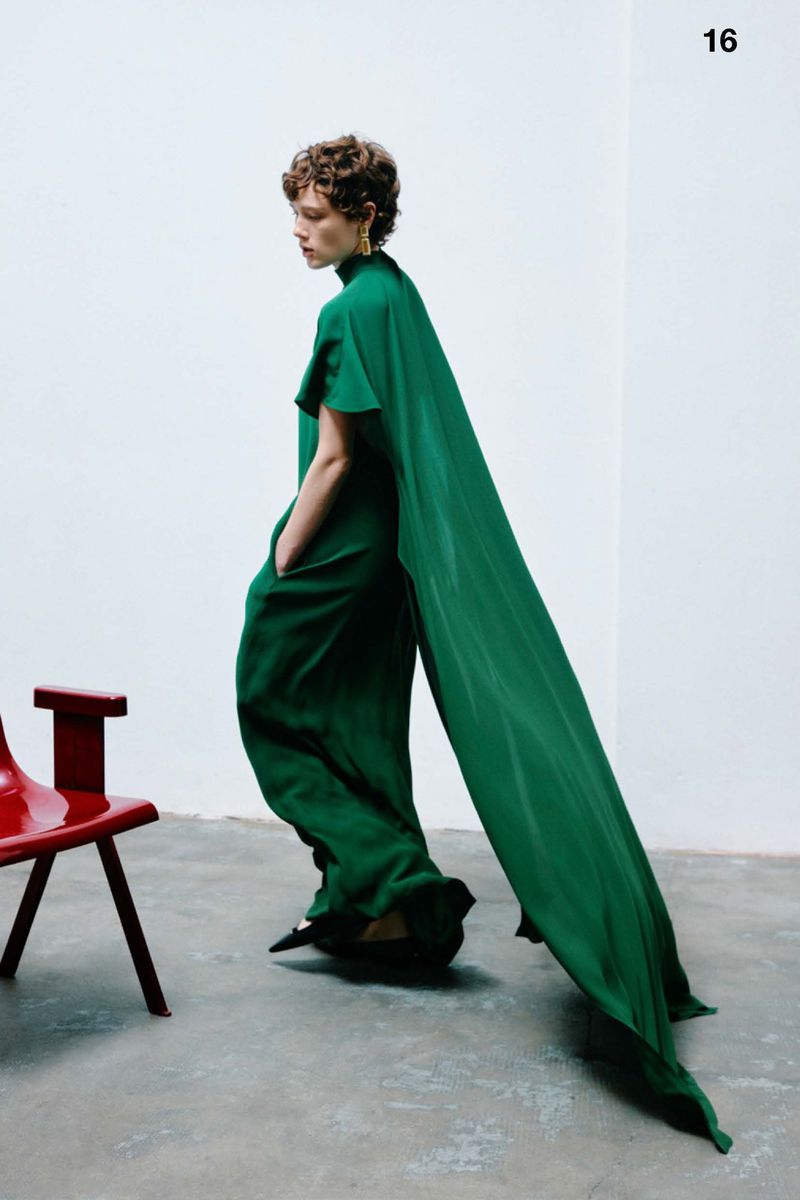 Fashionable woman in a green jumpsuit with a long cape, walking in a photo studio with a red chair, creating a striking editorial image.