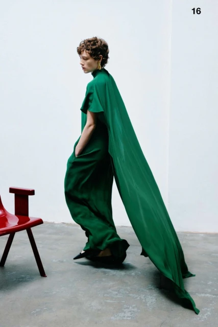 Fashionable woman in a green jumpsuit with a long cape, walking in a photo studio with a red chair, creating a striking editorial image.