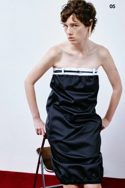 A stylish woman poses in a black dress against a white backdrop, leaning on a folding chair, with red flooring adding a pop of color.