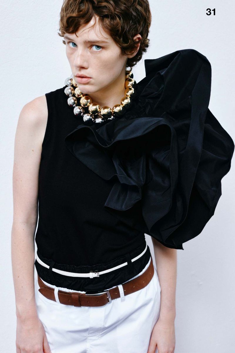 Fashion editorial shot of a young adult woman with short brown hair wearing a black top, statement necklace and white pants in a studio.