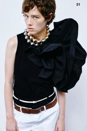 Fashion editorial shot of a young adult woman with short brown hair wearing a black top, statement necklace and white pants in a studio.