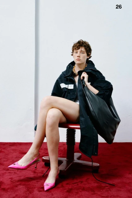 Fashion shot of a female-presenting model in a black jacket, gold necklace, and pink heels, seated on a red-cushioned chair.