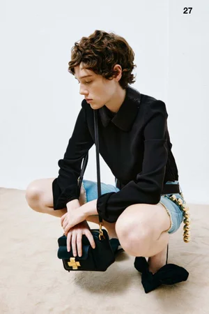 Model crouching against a seamless white background wearing a black collared top, denim shorts, and holding a dark handbag.