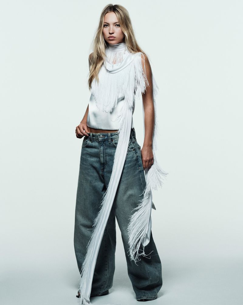 Full-length shot of an adult female fashion model wearing an off-white fringed top and blue jeans in a photo studio setting.