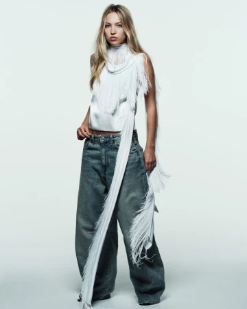 Full-length shot of an adult female fashion model wearing an off-white fringed top and blue jeans in a photo studio setting.