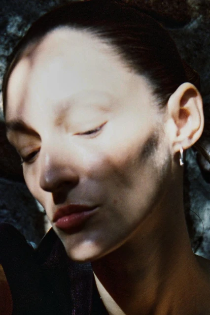 Close-up portrait of a woman with fair skin, closed eyes, and an earring, bathed in dappled light against a muted, dark background.
