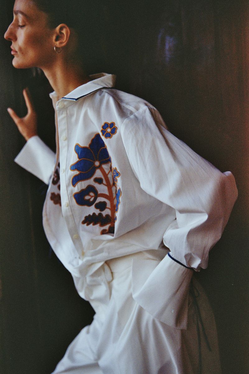 Adult female in white button-down shirt with blue and brown floral design, posing indoors against a dark wall in soft, diffused light.