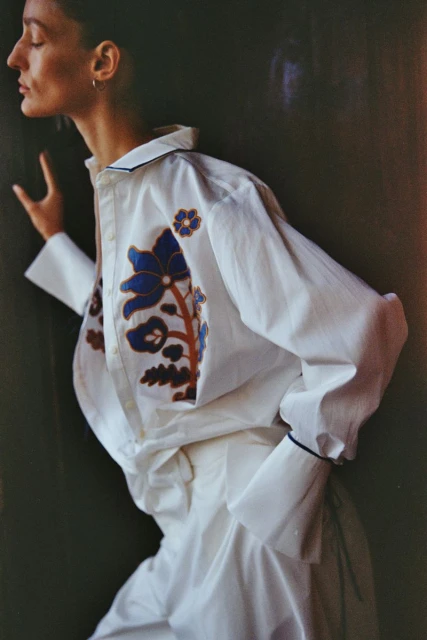 Adult female in white button-down shirt with blue and brown floral design, posing indoors against a dark wall in soft, diffused light.