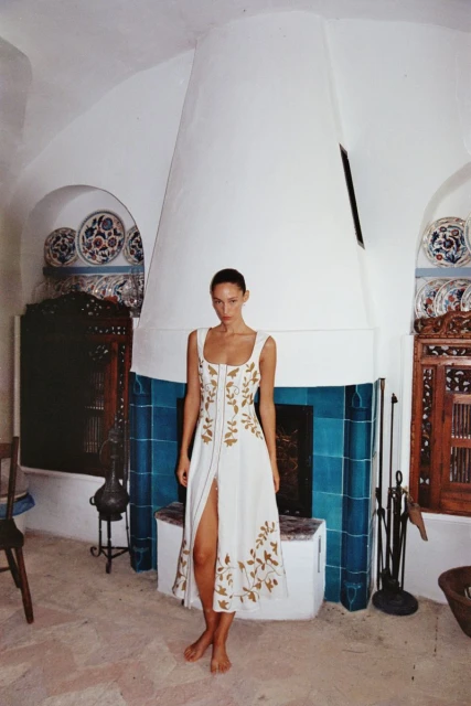 A woman poses in a white dress with gold leaf patterns in an airy, white-walled interior with blue-tiled accents and a white floor.