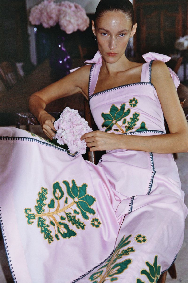 A woman wearing a pink dress with green floral embroidery sits indoors, holding a pink hydrangea with a vase of flowers in the background.