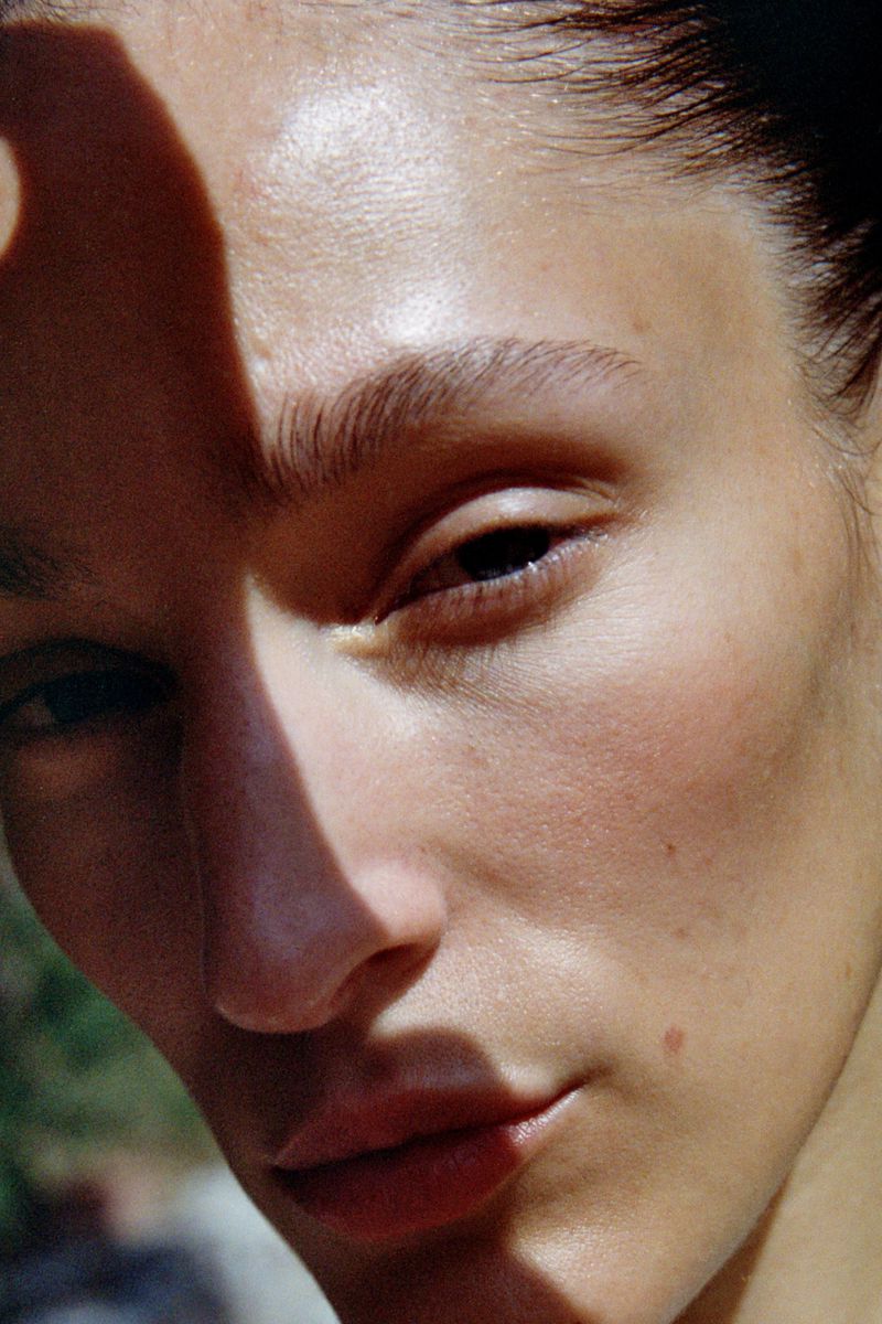 Close-up portrait of a young adult woman's face, with half her face shadowed, showcasing natural skin texture and minimalistic beauty look.