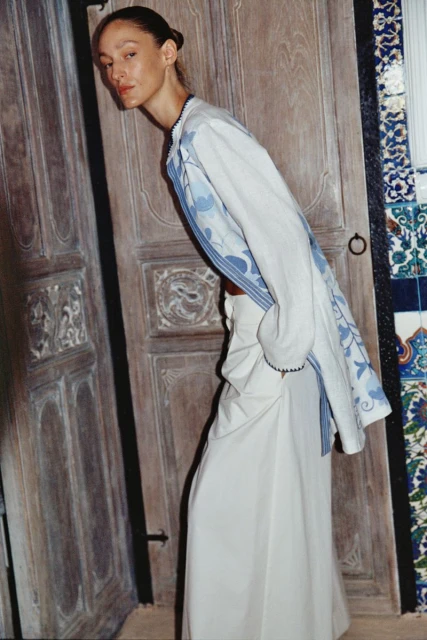 Fashionable adult female model posing in a white linen dress and floral print jacket near an ornate wooden door inside a studio.